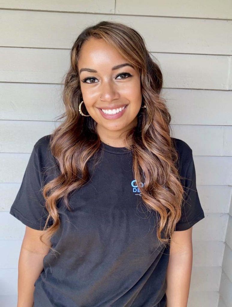 The image shows a smiling woman with long brown hair wearing a black t-shirt and standing against a beige wall.