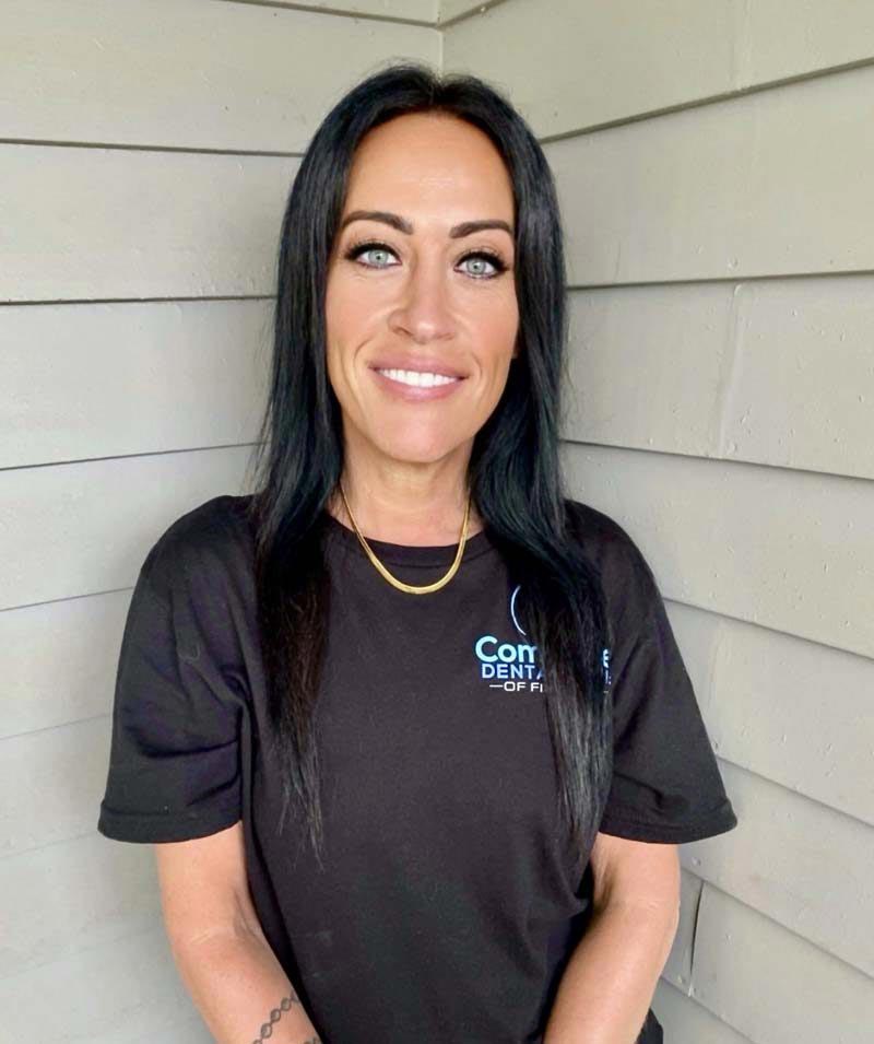 The image shows a woman posing against a wooden wall with a neutral expression, wearing a dark top with a logo on it, and a necklace around her neck.