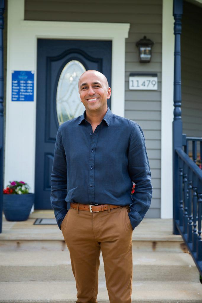 The image shows a man standing on a porch in front of a house, smiling at the camera.