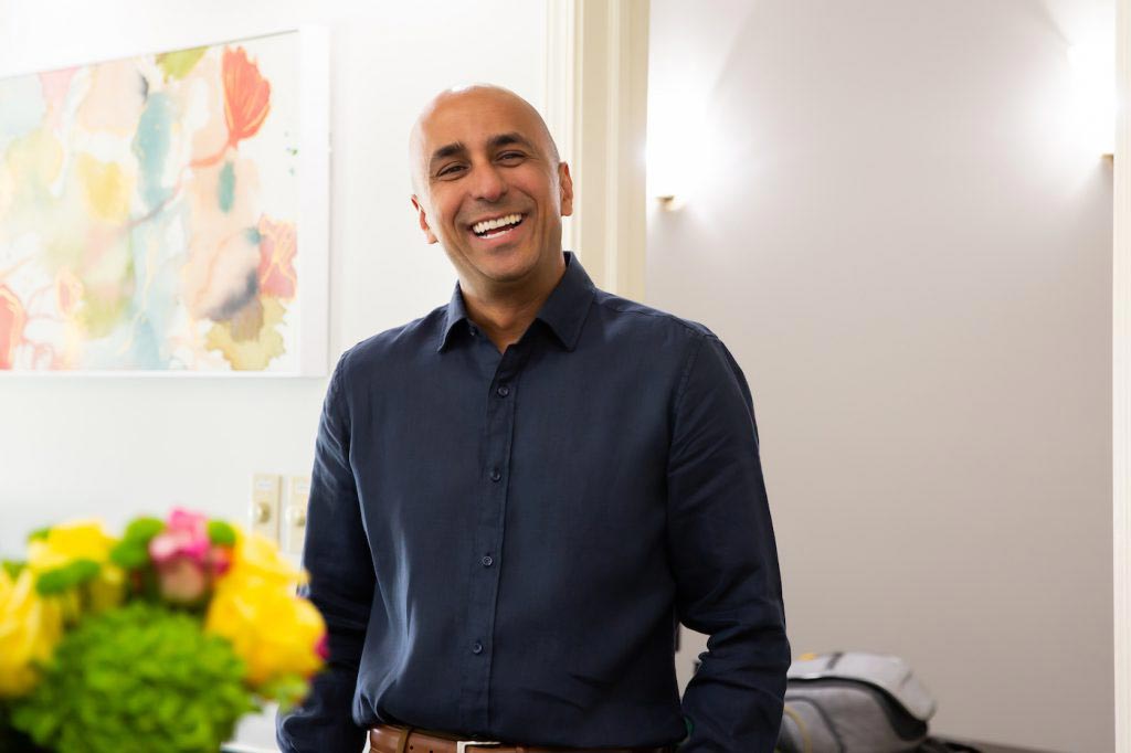 The image shows a man standing indoors, smiling at the camera. He has dark hair, a beard, and is wearing a dark shirt. There s a wall-mounted light fixture above him, and he appears to be in a room with decorative elements and potted plants.