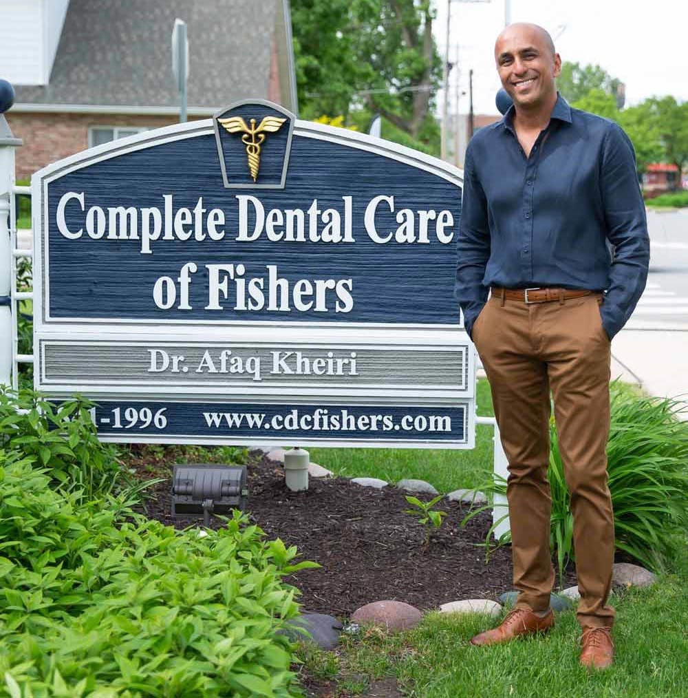 The image features a man standing in front of a sign for Complete Dental Care of Fishers, smiling at the camera. He is wearing a dark shirt with rolled-up sleeves, light brown pants, and brown shoes. Behind him is a green lawn with some plants and a building with a brick facade.