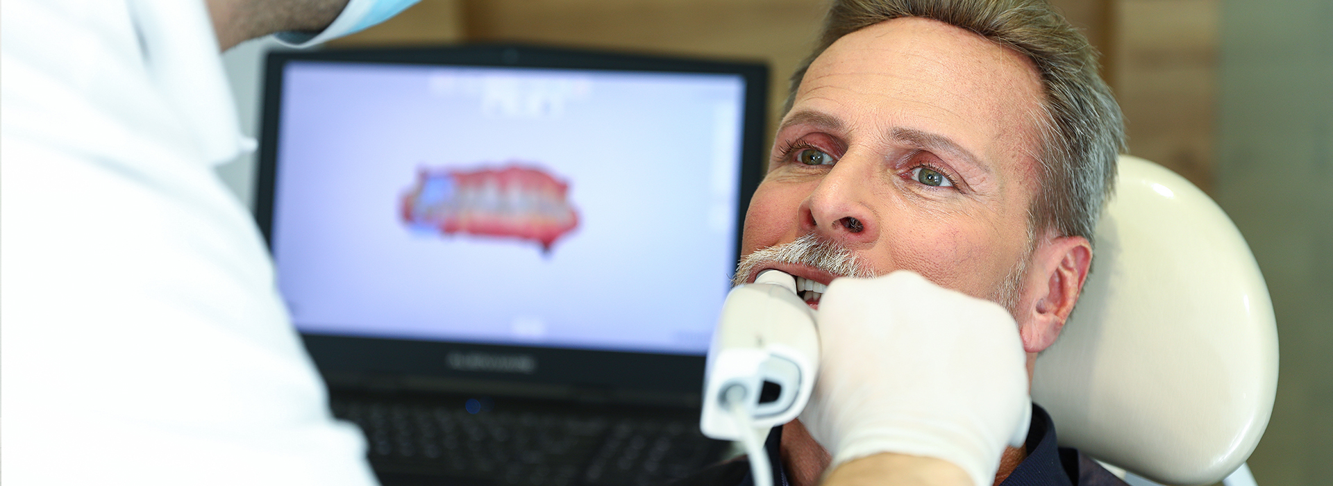 The image shows a man receiving dental treatment, with a dentist performing an examination using a digital device while another person looks on.