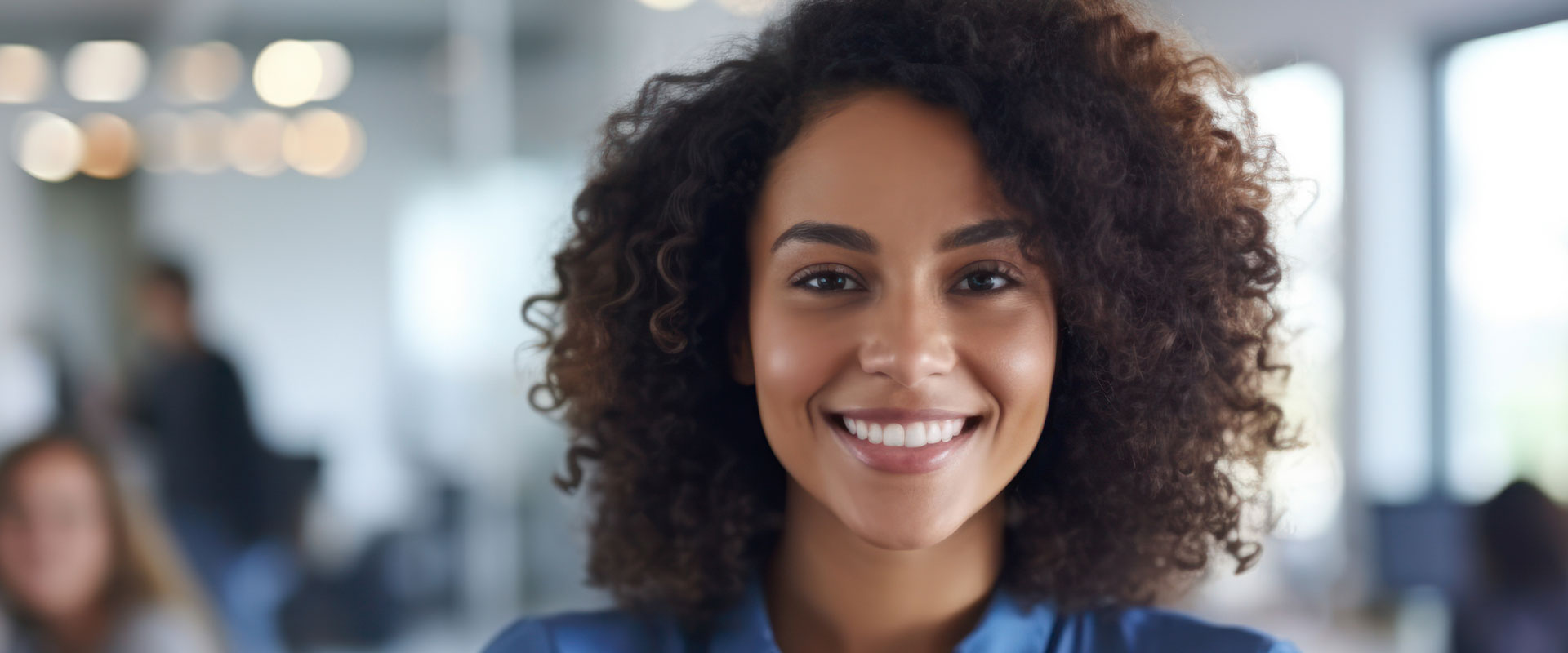 The image shows a woman with short curly hair, wearing a dark top, smiling at the camera. She appears to be in an office environment with other people in the background.