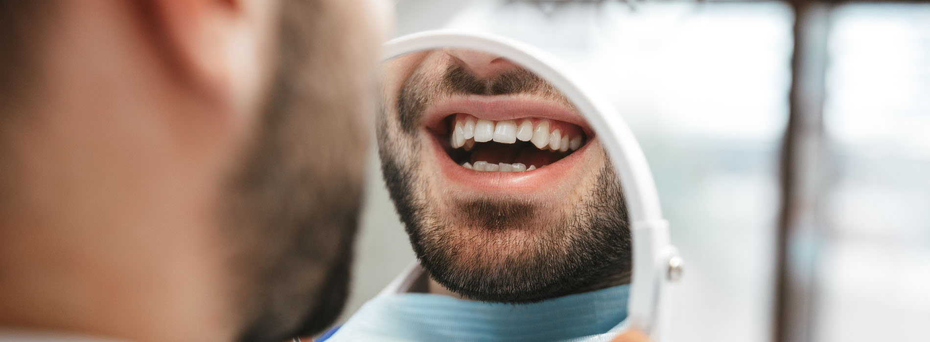 The image shows a close-up of a man with a beard looking at his teeth while sitting in a dental chair, with a dental professional standing behind him.