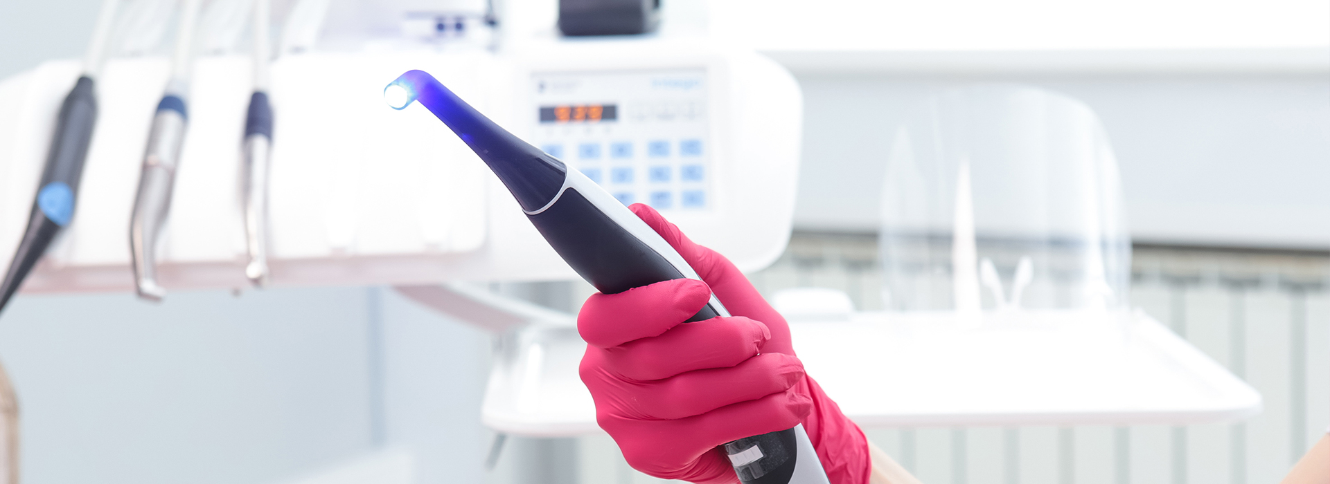 The image shows a person wearing a red glove holding a blue device with a black handle, possibly an electronic tool or gadget, in front of a white wall with various equipment and a refrigerator in the background.