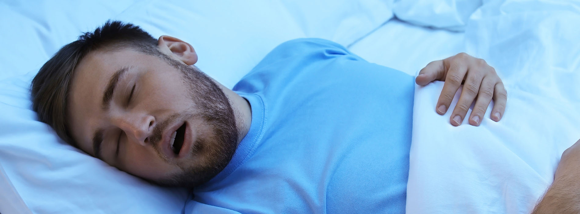 A man lying down with his head on a pillow, wearing a blue shirt, against a background that appears to be a bed.
