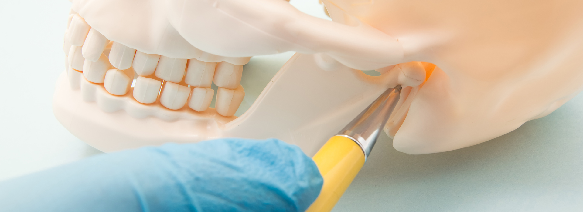 A cropped image showing a close-up of a dental model with a tooth being worked on by a dental tool, with a blue cloth background and a hand holding a small yellow object.