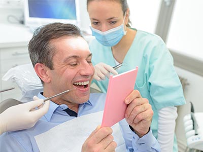 A man sits in a dental chair with a cardboard sign on his chest, while a dentist and hygienist look at him, smiling at the camera.