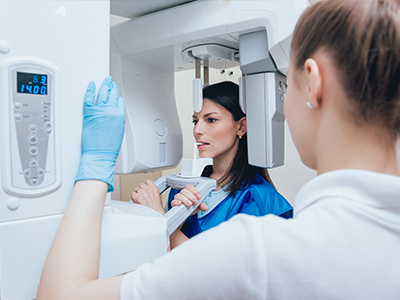 Two individuals are present in the image, with one person standing behind a large medical imaging machine, possibly an MRI or CT scanner, while another person stands in front of it, seemingly observing the equipment or receiving an explanation about its operation. The setting appears to be a clinical environment, and both individuals are wearing protective gloves, suggesting adherence to hygiene protocols.