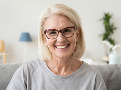 The image shows a woman with blonde hair wearing glasses, smiling at the camera, against a blurred indoor background that includes furniture and decorative items.