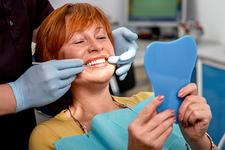 A woman sitting in a dental chair with a blue mouthguard on her teeth, smiling at the camera while holding up a mirror with a smiley face drawn on it.