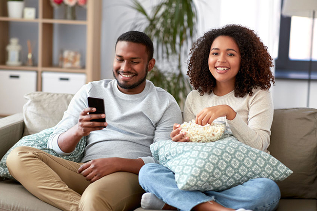 A man and woman sitting on a couch, smiling and enjoying a movie together, with popcorn visible between them.