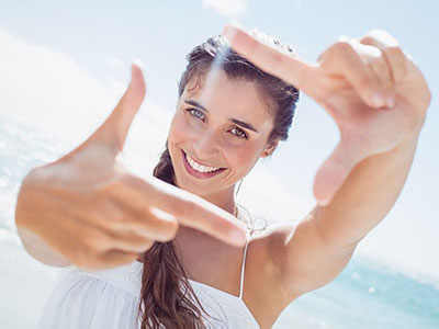 A young woman with long hair, wearing a white top, is holding her hand up to her face, making a peace sign with her fingers, and smiling at the camera while standing on a beach.