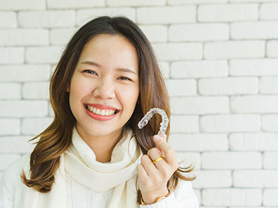 The image features a young woman holding up a toothbrush with a smile on her face, standing in front of a brick wall.